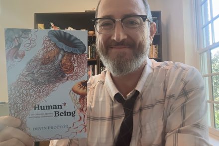 A man in a white shirt and black tie holds a book called "Human*Being"